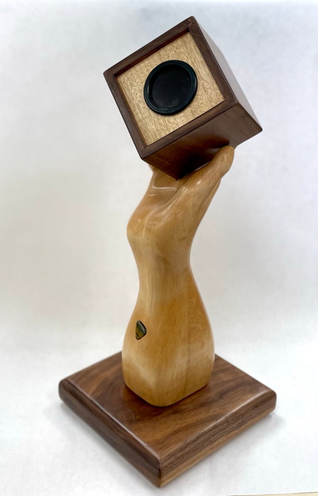 A life-sized sculpted arm and hand in maple grasps a walnut cube that encloses a Bluetooth enabled speaker. The cube is removable for charging. The sculpture or 18” tall on a 7” square walnut base